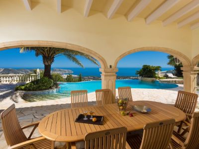 Expansive covered terrace featuring a wooden dining table and chairs, framed by stone arches revealing panoramic Mediterranean sea and coastal views with an infinity pool.