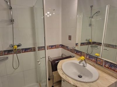 Functional bathroom featuring a glass shower cabin, vanity with light marble countertop, and an illuminated mirror. White tiles with a decorative Mediterranean border.