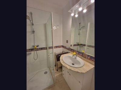 Functional bathroom featuring a walk-in shower with a glass screen, a basin set in a light marble-patterned countertop, and a white vanity. White wall tiles are accented with a decorative Mediterranean-style border.