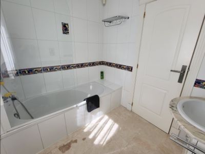 Functional bathroom with a white bathtub, glass screen, and walls tiled in white with a decorative border. Features a marble-topped sink and natural light.
