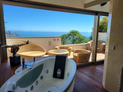 Indoor jacuzzi with wooden flooring opening onto a private terrace, furnished with rattan seating and offering panoramic Mediterranean sea views.
