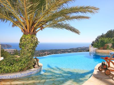 Infinity pool offering panoramic sea and coastal views, featuring an ivy-covered palm tree and a wooden bar cart on the stone sun terrace.