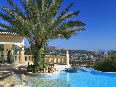 Infinity-style pool featuring a palm tree island and an integrated table, with panoramic coastal and mountain views under a clear sky.