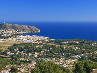 Panoramic view from a hillside, featuring a coastal town, a marina, a prominent mountain, and the deep blue Mediterranean Sea under a clear sky.
