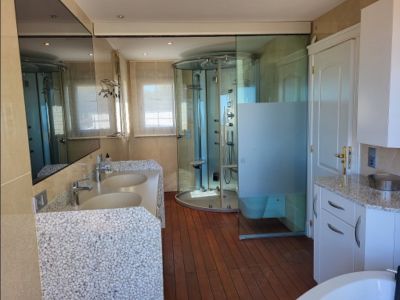 Spacious bathroom featuring a double vanity with pebble-textured base, a circular hydro-massage shower, and warm wooden flooring. Large mirror and windows with blinds are visible.