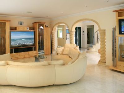 Spacious living room featuring a large curved cream sofa, light marble floors, and custom light wood entertainment units. Arched stone columns lead to the entrance and staircase.