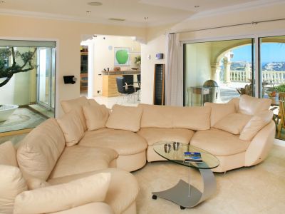 Spacious living room featuring a light sectional sofa, glass coffee table, and marble floors. Large windows offer views to a sea-facing terrace and an internal olive tree courtyard.