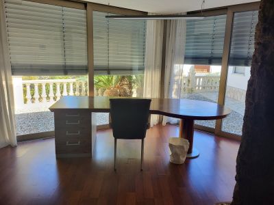 Spacious private office featuring a custom wooden desk and floor-to-ceiling windows with views of a gravel patio and Mediterranean garden. Dark wood flooring and modern lighting.
