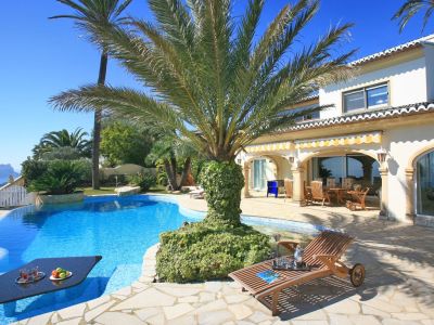 The uniquely designed pool features an in-water table for refreshments, surrounded by a stone terrace and palm trees, with distant sea views.
