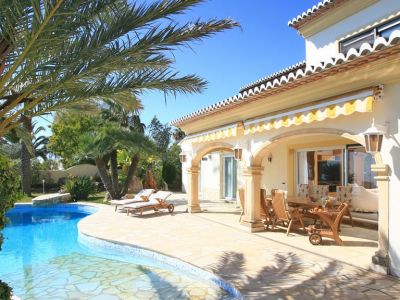 The villa's exterior features a freeform swimming pool and a spacious stone-paved terrace leading to a covered naya with a wooden dining set, all under striped awnings.