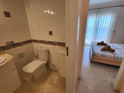 View from a tiled bathroom with a toilet and sink, looking into a bright bedroom featuring twin beds with striped linens and a large window with sheer curtains.