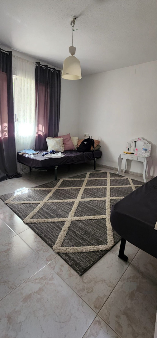 Bedroom featuring light tiled flooring, a geometric grey and beige rug, and a single bed by the window with dark curtains. A small white dressing table is also visible.