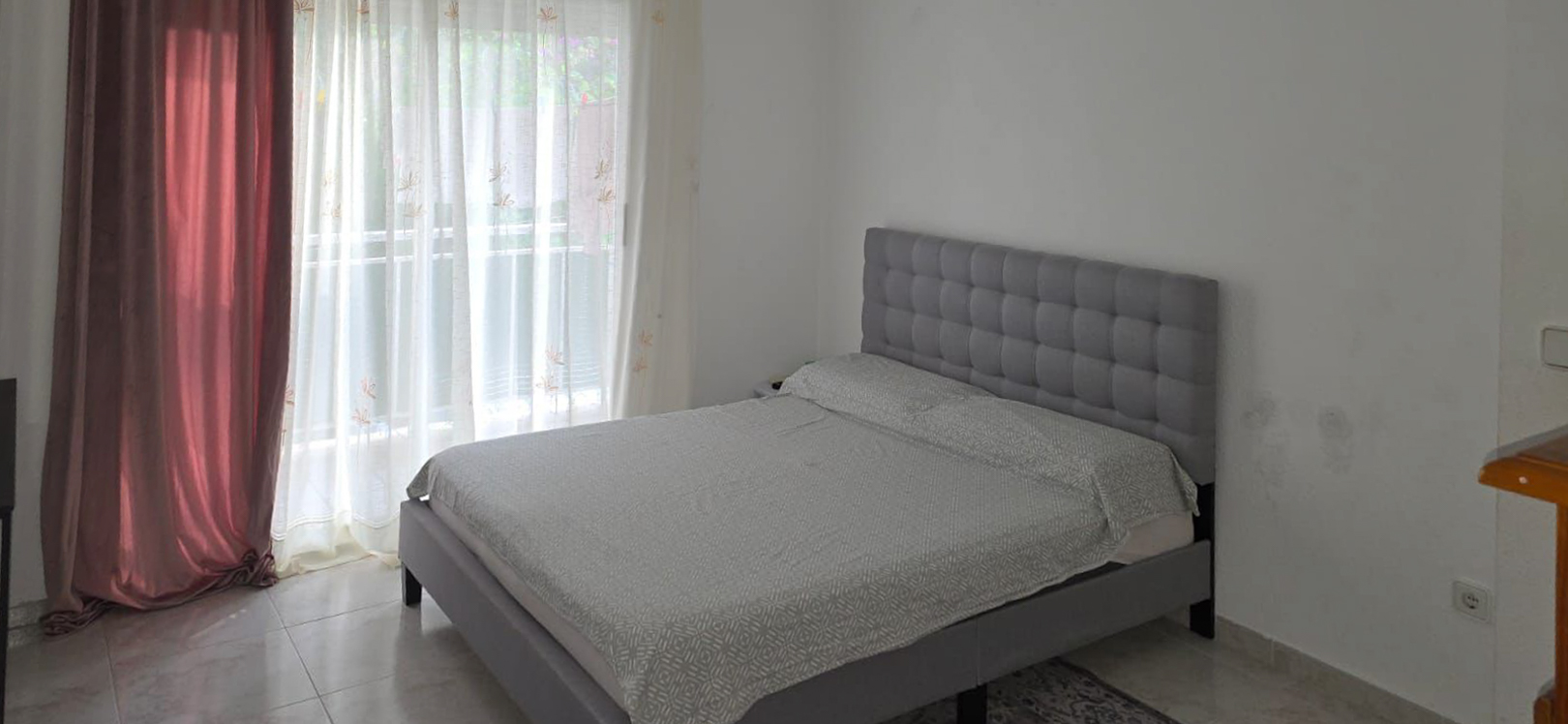 Bedroom with a grey upholstered bed and tufted headboard, complemented by light patterned bedding. Natural light enters through the large window, featuring sheer and dark red curtains, over tiled flooring.