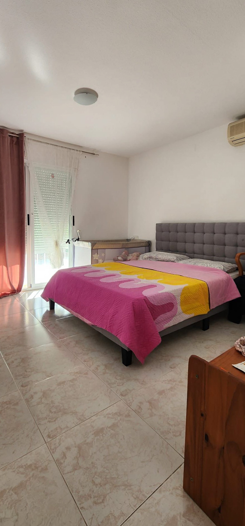 Bedroom with a grey upholstered bed, light tiled floor, natural light from a window with roller shutter, and a baby crib. Equipped with wall-mounted air conditioning.