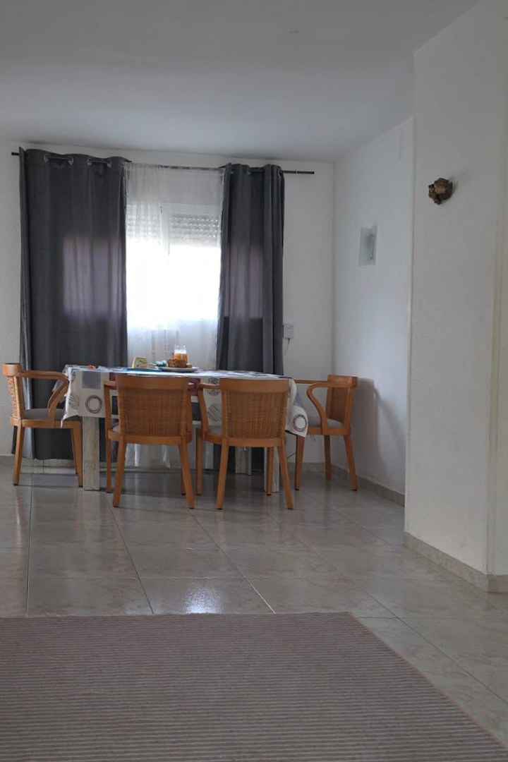 Bright dining area featuring a wooden table with woven chairs, positioned by a window with grey curtains. Light-colored tiled flooring extends throughout, with a striped rug in the foreground.