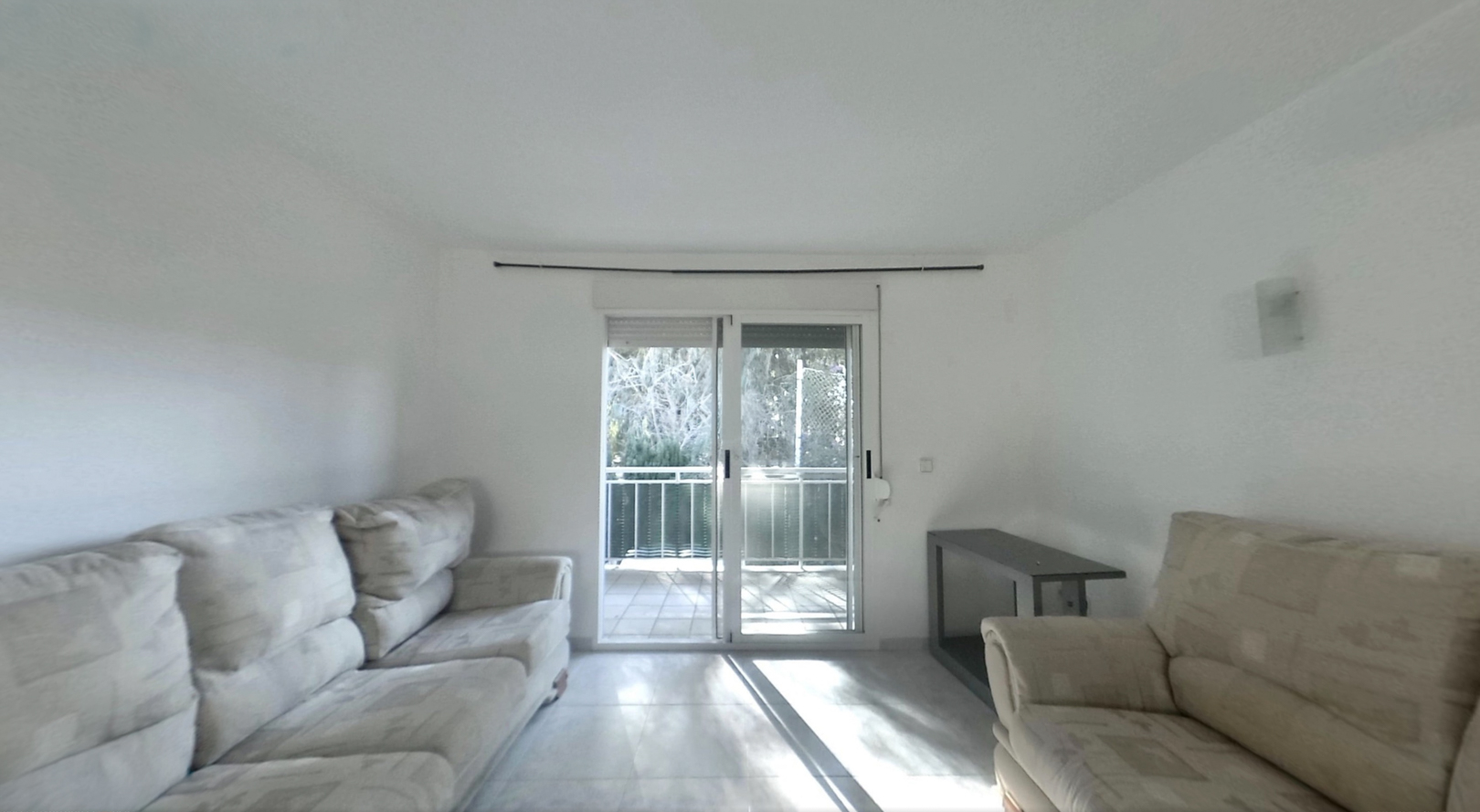 Bright living room featuring light-colored sofas, tiled flooring, and sliding glass doors that open to a balcony, allowing natural light to illuminate the space.