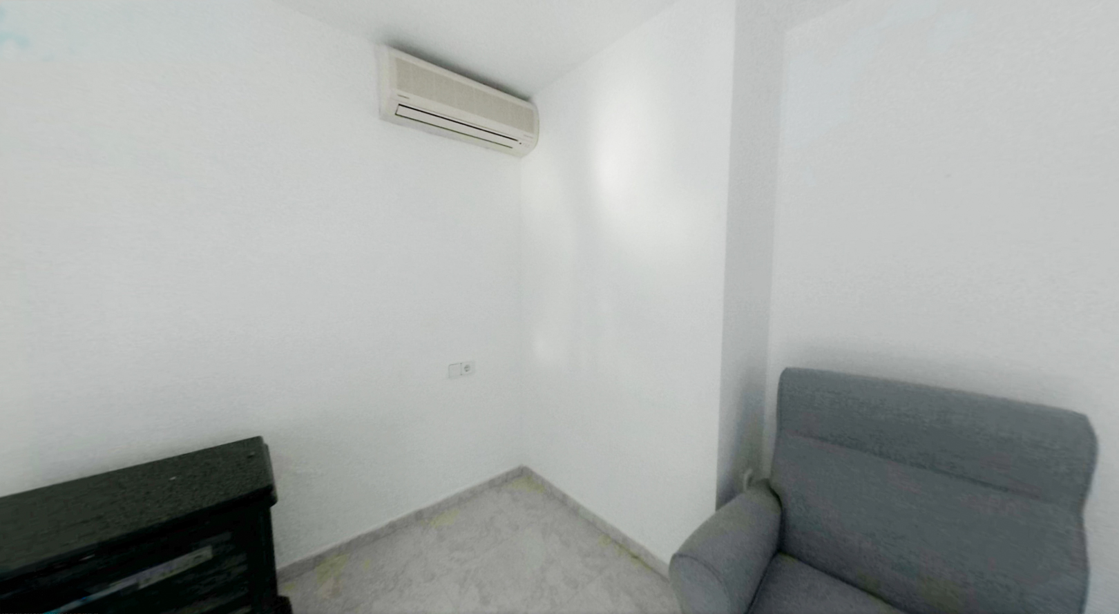 Corner of a room featuring white walls and light tiled flooring. An air conditioning split unit is mounted, with a power outlet below, beside a grey armchair and dark furniture.
