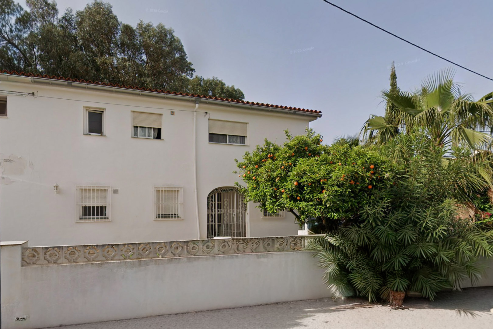 Exterior view of the white villa featuring a red tiled roof, windows with security bars, and an arched entrance. A vibrant orange tree and lush greenery adorn the front garden.