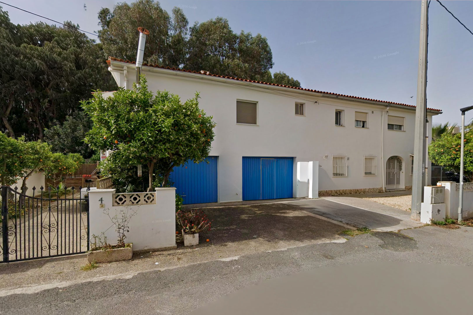 Front exterior of the semi-detached villa, featuring a white facade, two distinctive blue garage doors, and a mature orange tree by the wrought-iron entrance gate.