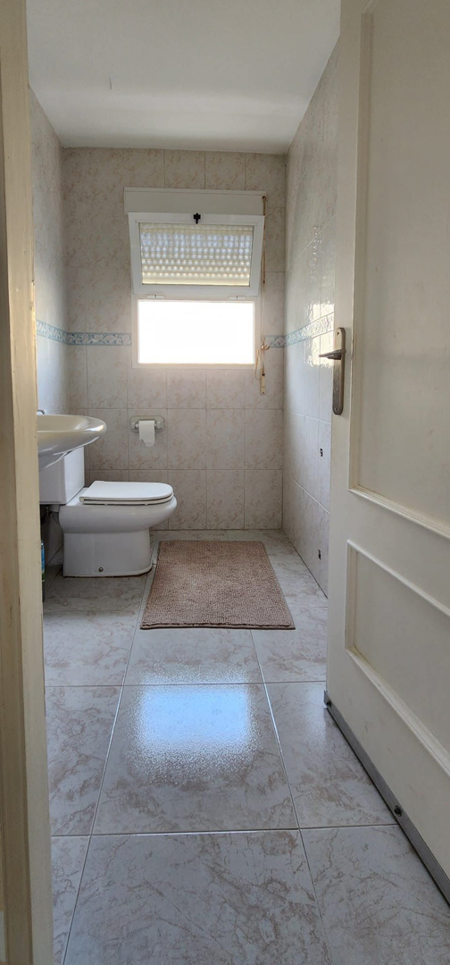 Functional bathroom featuring a toilet, sink, and a window with blinds. It has light cream wall and floor tiles with a blue decorative border and a bath mat.