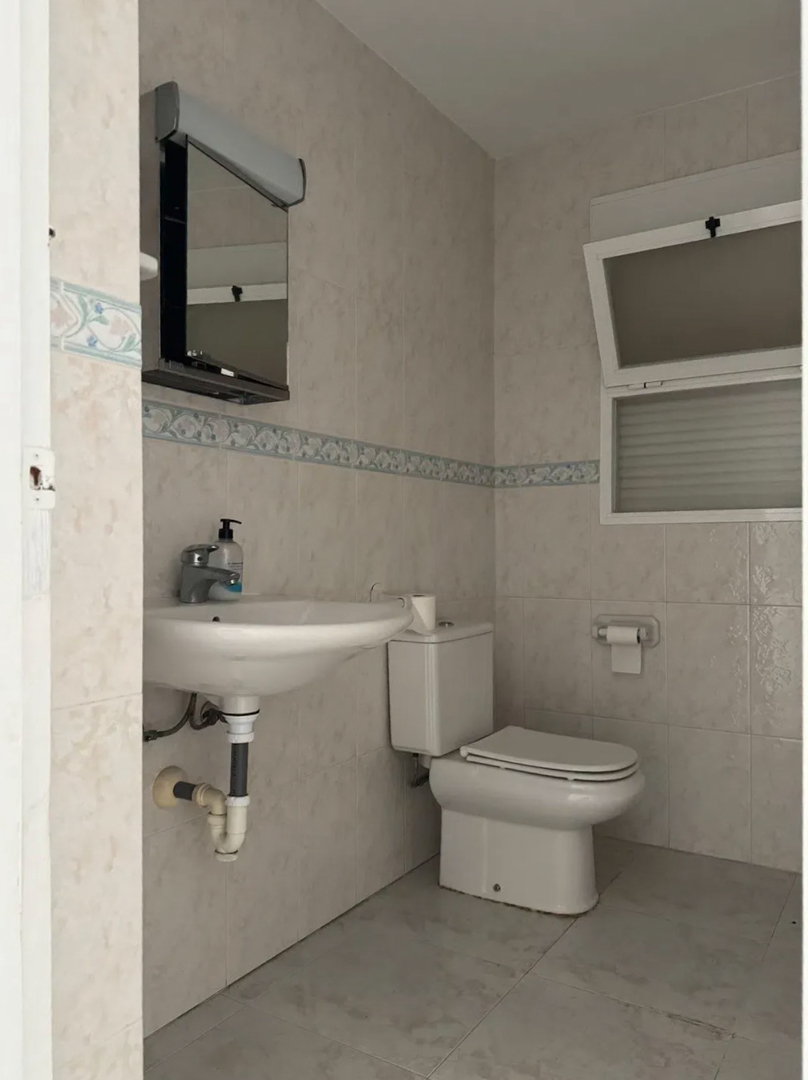 Functional bathroom featuring light-toned wall tiles with a decorative border, a wall-mounted sink, toilet, and an illuminated mirror. A tilt-and-turn window provides natural light and ventilation.