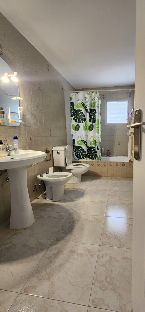 Functional bathroom with light marble-effect floor tiles and beige wall tiles. Features a pedestal sink, toilet, bidet, and bathtub with a leaf-patterned curtain and frosted window.