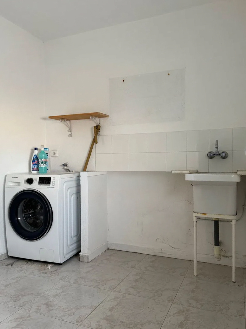 Functional utility room featuring a washing machine, a practical utility sink with a tiled backsplash, and light-colored floor tiles. A wooden shelf is visible.