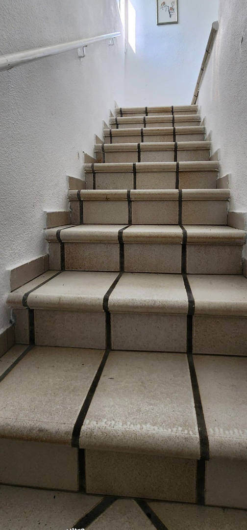 Interior staircase featuring light-colored tiled steps with dark grout, textured white walls, and simple handrails. Natural light illuminates the upper landing.