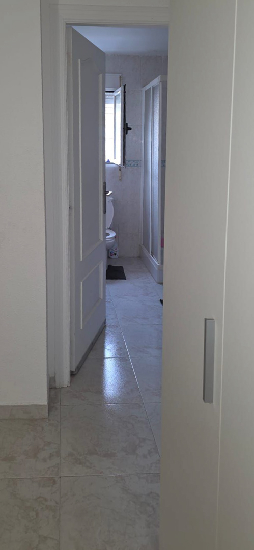 Partial view into a bathroom featuring light-toned floor tiles, a toilet, and a window providing natural light. Includes a shower enclosure and decorative wall tiling.