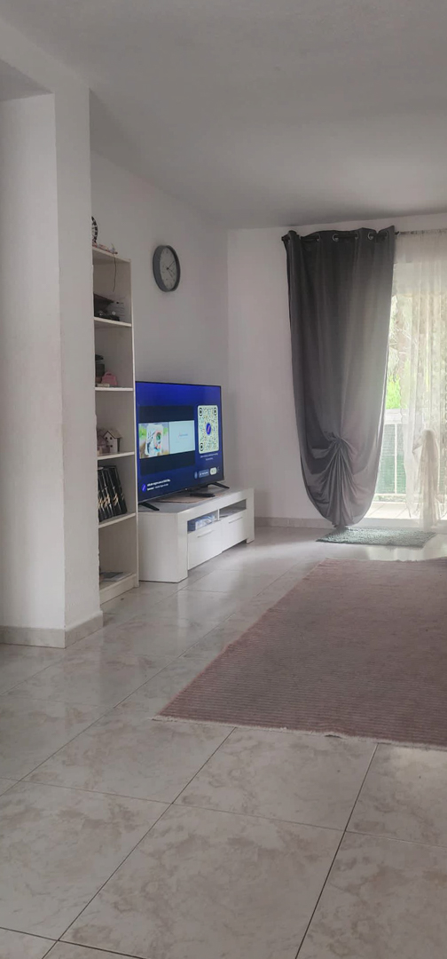 Partial view of the living room featuring a TV on a white console, a shelving unit, and a glass door with grey and sheer curtains opening to a terrace, set on light tiled flooring with a rug.