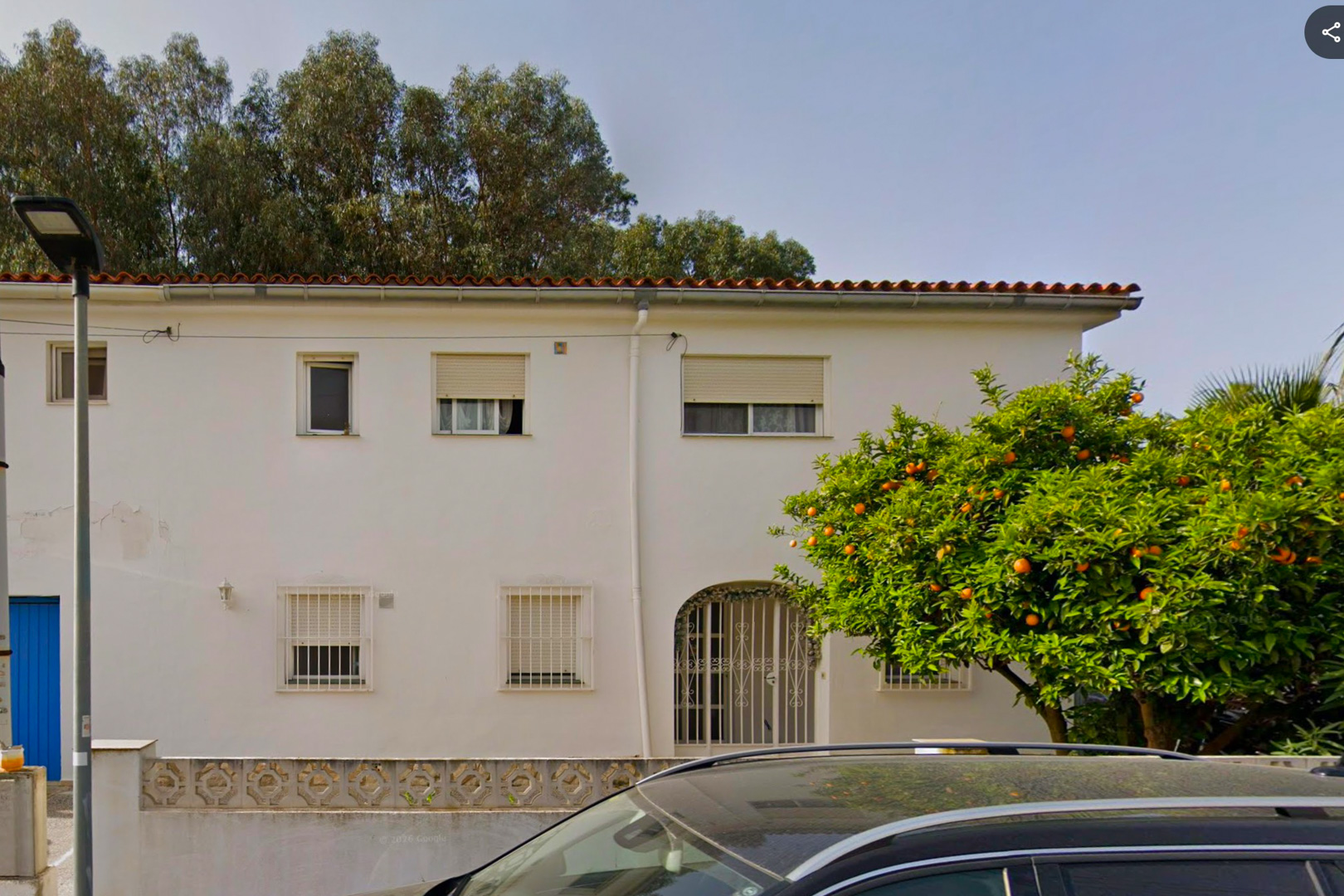 The bright white facade of the two-story villa features a traditional tiled roof, an arched entrance with an ornate gate, and windows with security bars. A mature orange tree, heavy with fruit, stands prominently.
