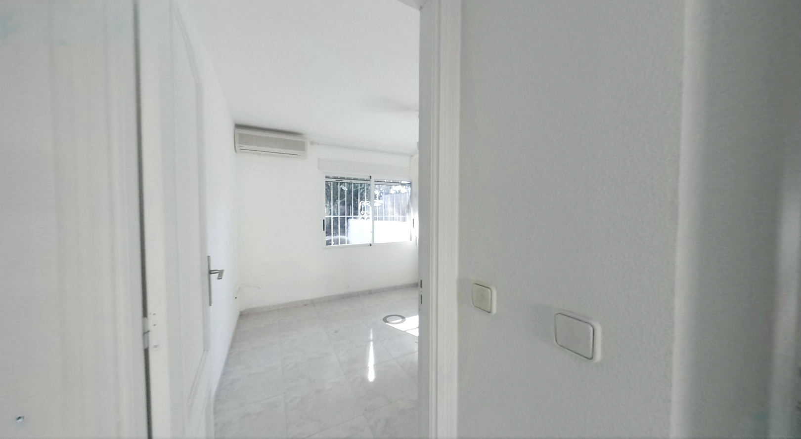 View into an unfurnished room featuring light tiled flooring, white walls, and an air conditioning unit. A window with security bars provides natural light.