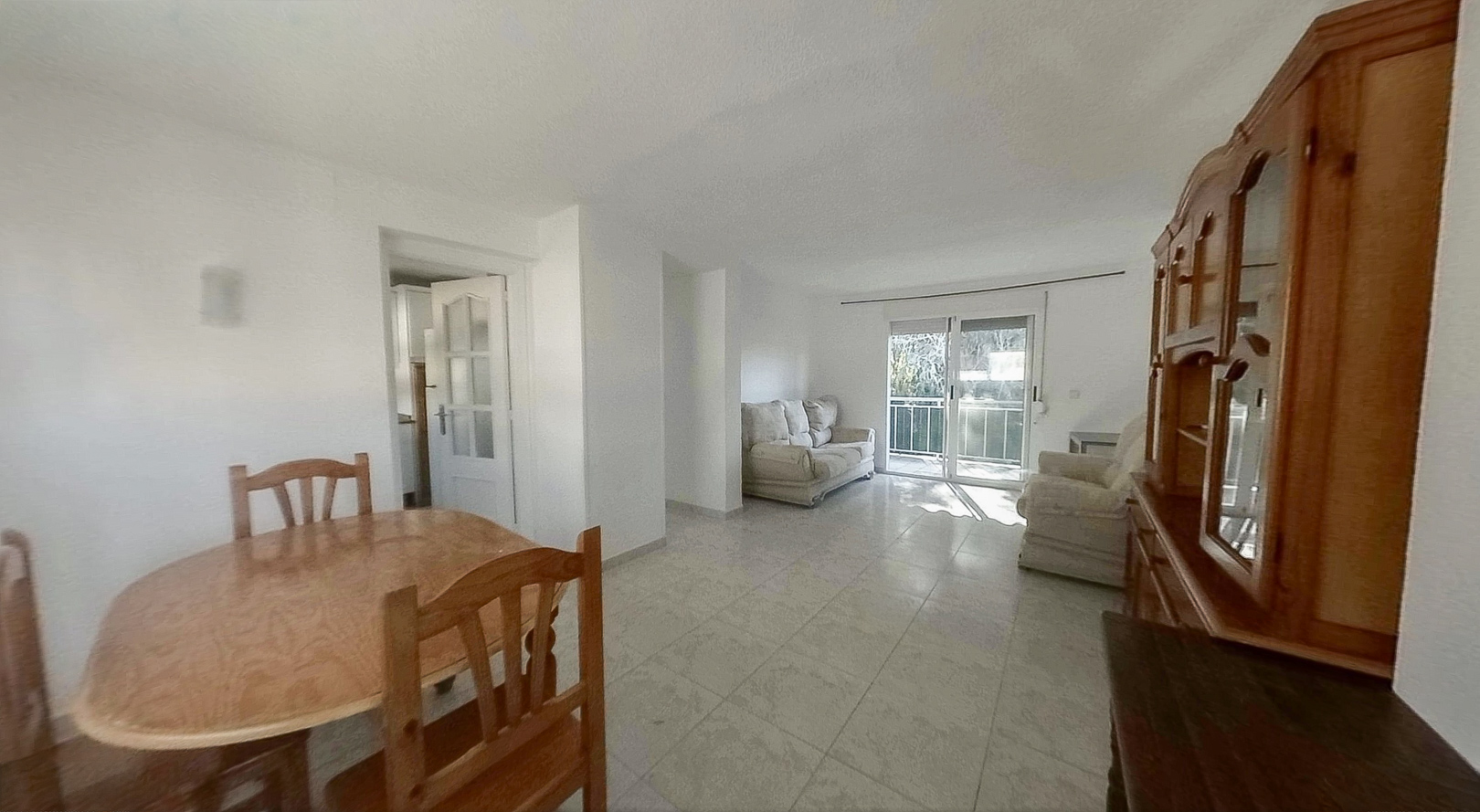 View of the bright living-dining area featuring an oval wooden table, tiled floors, and a large wooden display cabinet. Sliding glass doors open to a balcony, filling the space with natural light.