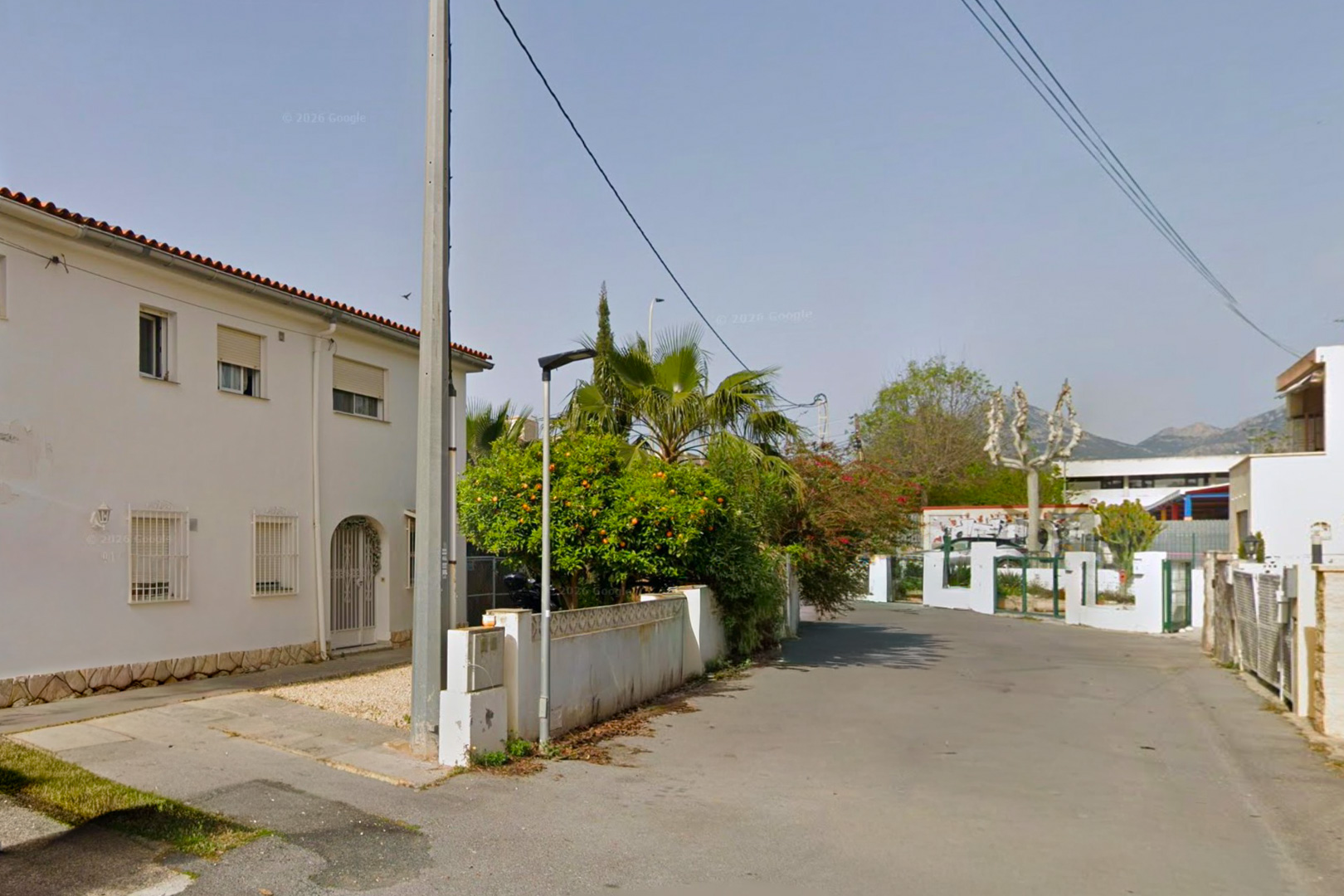 View of the property's two-story white facade with red roof tiles and security bars, featuring a gravel entrance path and a perimeter wall with lush vegetation including an orange tree.