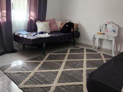 Bedroom featuring light tiled flooring, a grey geometric rug, and a daybed. Natural light enters through a window with dark curtains, complemented by a small white dressing table.