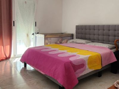 Bright bedroom featuring a double bed with a grey upholstered headboard, tiled flooring, and direct outdoor access via a glass door. Includes air conditioning.