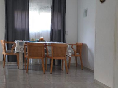 Bright dining area featuring a wooden table with woven chairs, positioned by a window with grey curtains. Light-colored tiled flooring extends throughout, with a striped rug in the foreground.