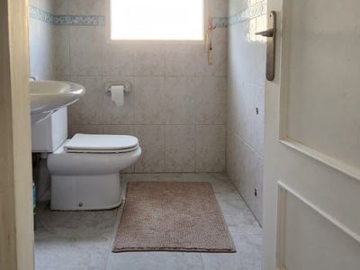Compact bathroom featuring a toilet, wall-mounted sink, and natural light from a window with blinds. Tiled walls and floor.