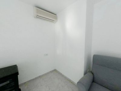 Corner of a room featuring white walls and light tiled flooring. An air conditioning split unit is mounted, with a power outlet below, beside a grey armchair and dark furniture.