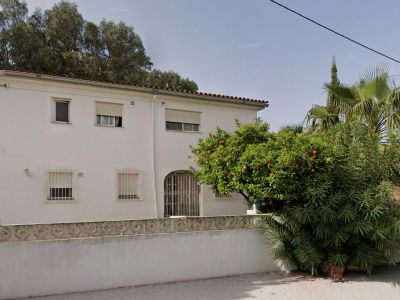 Exterior view of the white villa featuring a red tiled roof, windows with security bars, and an arched entrance. A vibrant orange tree and lush greenery adorn the front garden.