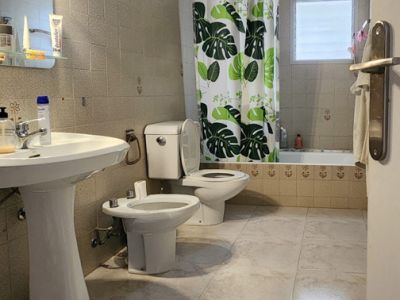 Functional bathroom with light marble-effect floor tiles and beige wall tiles. Features a pedestal sink, toilet, bidet, and bathtub with a leaf-patterned curtain and frosted window.
