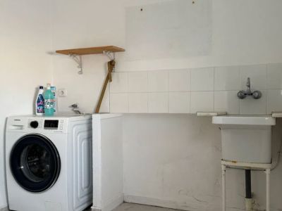 Functional utility room featuring a modern washing machine, a basic utility sink, and a white tiled backsplash. Light-colored floor tiles and a simple wooden shelf are visible.