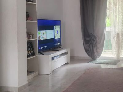 Partial view of the living room featuring a TV on a white console, a shelving unit, and a glass door with grey and sheer curtains opening to a terrace, set on light tiled flooring with a rug.