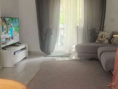 Spacious living room with natural light, featuring tiled floors, a comfortable sectional sofa, and direct access to a terrace through glass doors with elegant curtains.