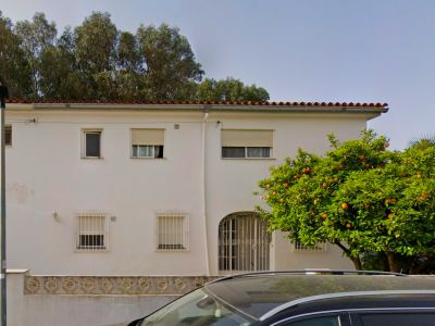 The bright white facade of the two-story villa features a traditional tiled roof, an arched entrance with an ornate gate, and windows with security bars. A mature orange tree, heavy with fruit, stands prominently.