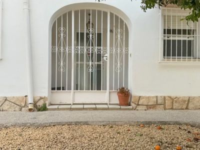 The villa's main entrance features a secure arched metal gate, leading to a glass door. A gravel garden with fallen fruit and an overhanging tree frames the facade.