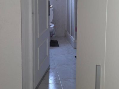 View into a bathroom featuring light-colored tiled flooring and white wall tiles with a decorative border. Includes a toilet, shower cabin, and natural light from a window.