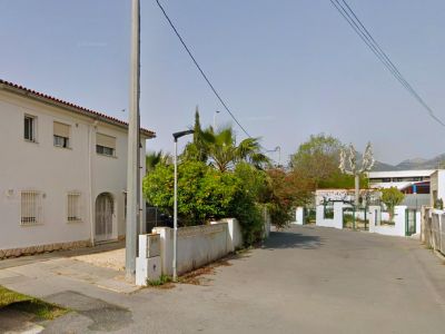 View of the property's two-story white facade with red roof tiles and security bars, featuring a gravel entrance path and a perimeter wall with lush vegetation including an orange tree.