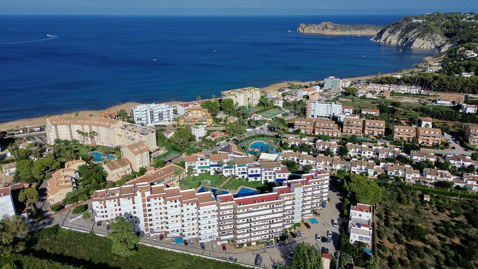 Aerial view of a coastal urbanization featuring multiple residential buildings, communal pools, and green areas, adjacent to a sandy beach and the Mediterranean Sea, with a distant rocky cape.