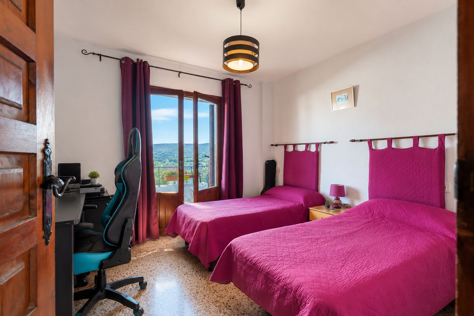 Bright bedroom featuring two single beds with fuchsia headboards and direct balcony access offering mountain views. Includes a desk area and terrazzo flooring.